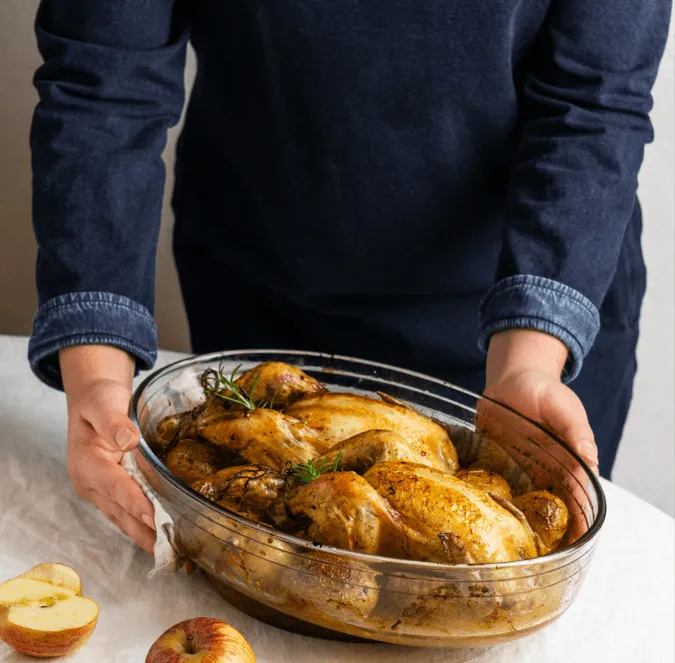 A person holding a glass dish with roasted whole chicken and potatoes, garnished with rosemary, placed on a white table with apples nearby