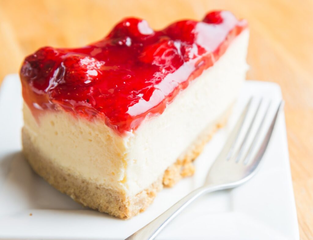 A slice of creamy cheesecake topped with glossy red strawberry glaze served on a white plate with a fork, placed on a wooden table.