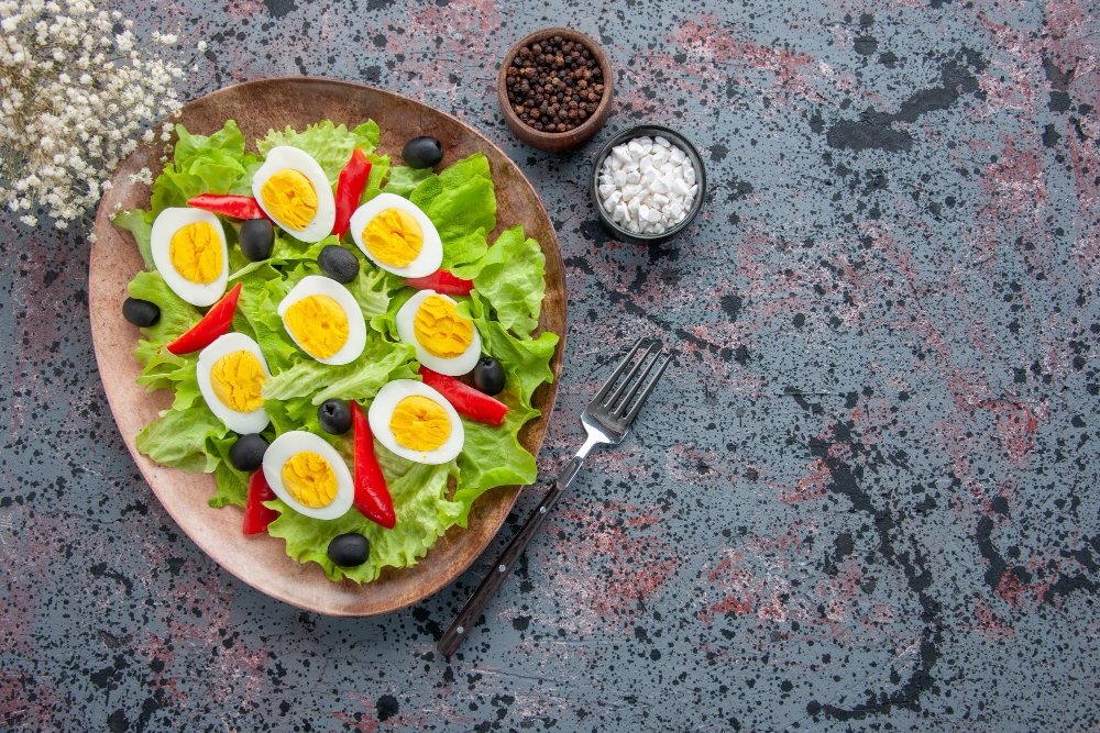 Healthy salad with lettuce, boiled eggs, black olives, and red bell peppers on a rustic plate, served with seasoning.