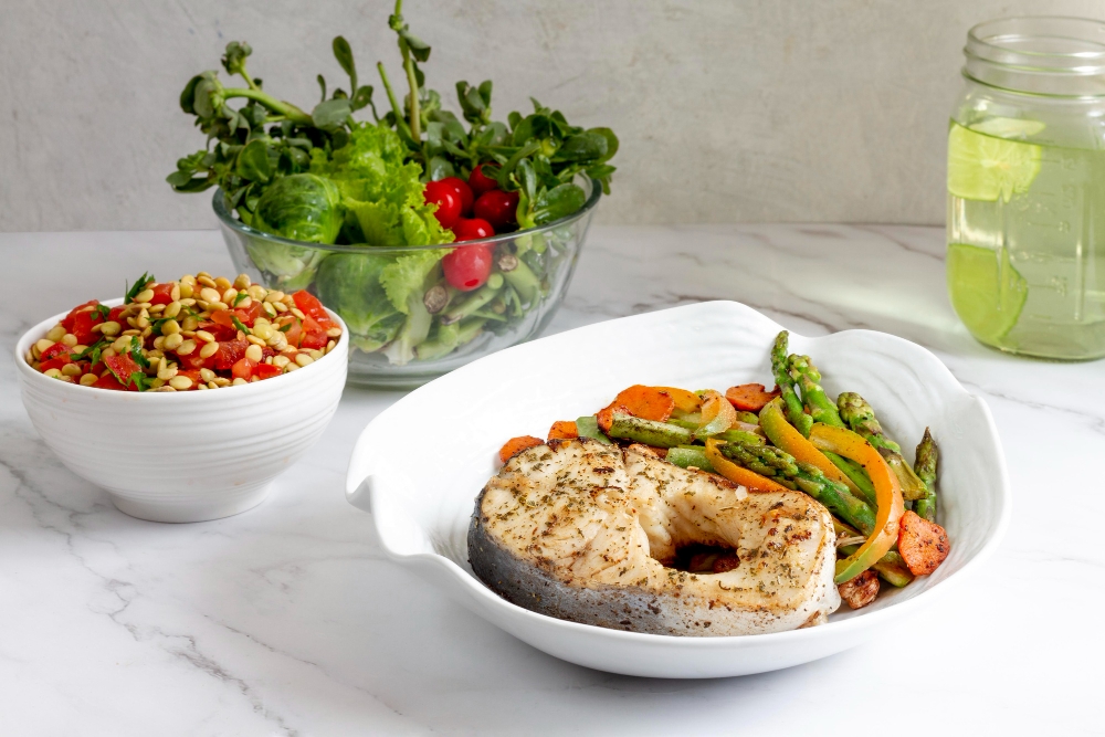 Grilled fish steak with sautéed vegetables, a fresh salad bowl, and a glass of lime water on a marble countertop.