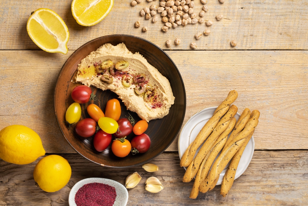 Hummus with olive oil, cherry tomatoes, lemon, garlic, and breadsticks on a wooden table.