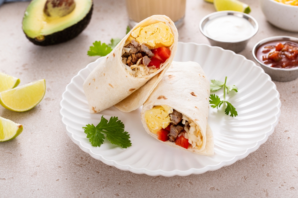 A close-up of two breakfast burritos filled with scrambled eggs, sausage, cheese, and diced red peppers on a white plate garnished with cilantro. Sides of salsa, sour cream, avocado, and lime wedges are in the background.