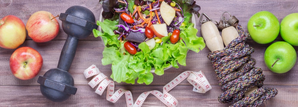 A healthy lifestyle flat lay with fresh apples, a colorful salad bowl, a dumbbell, a jump rope, and a measuring tape on a wooden background.