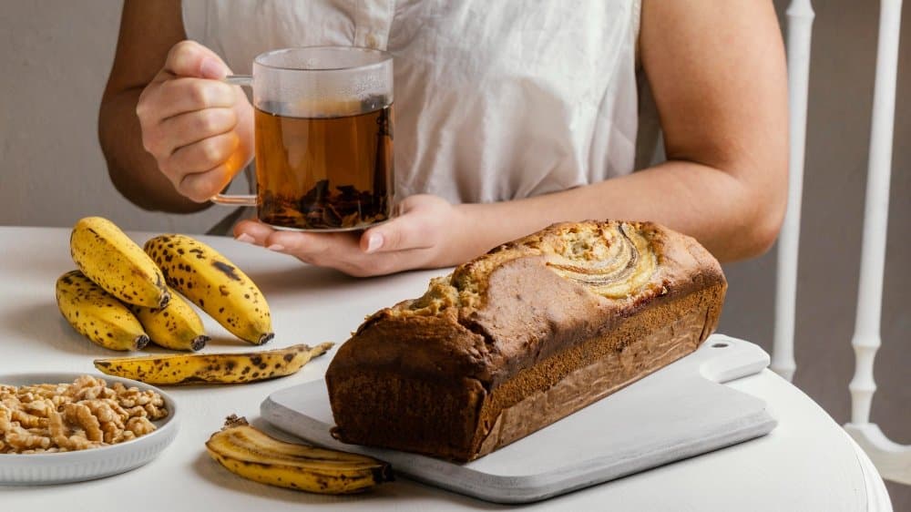 A freshly baked banana bread loaf on a white cutting board, surrounded by ripe bananas and walnuts, with a person holding a cup of tea.