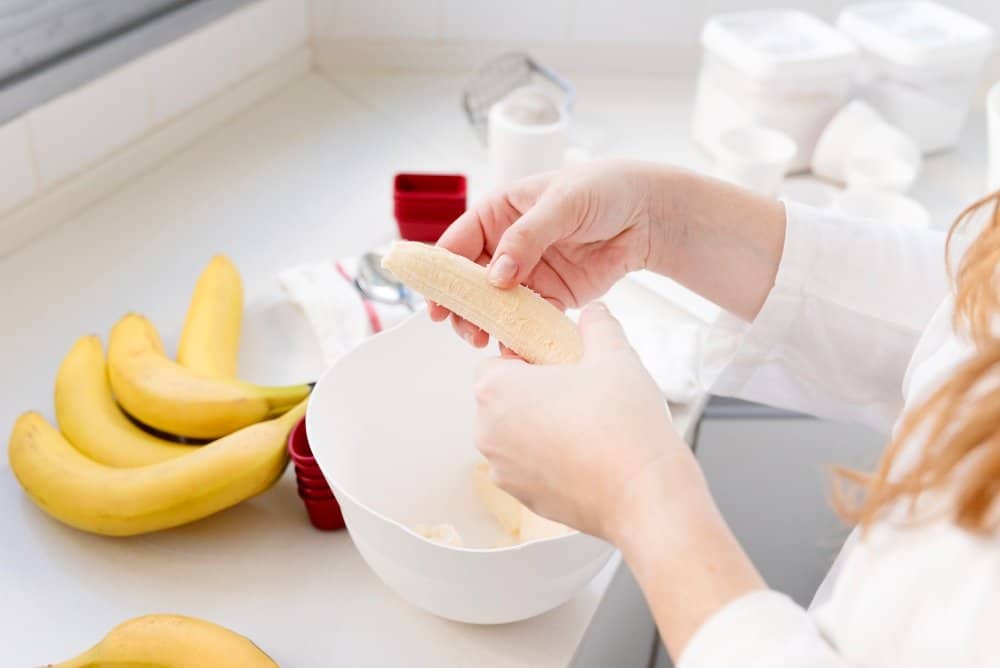 A person peeling a ripe banana into a mixing bowl in a bright kitchen, surrounded by fresh bananas and baking tools.