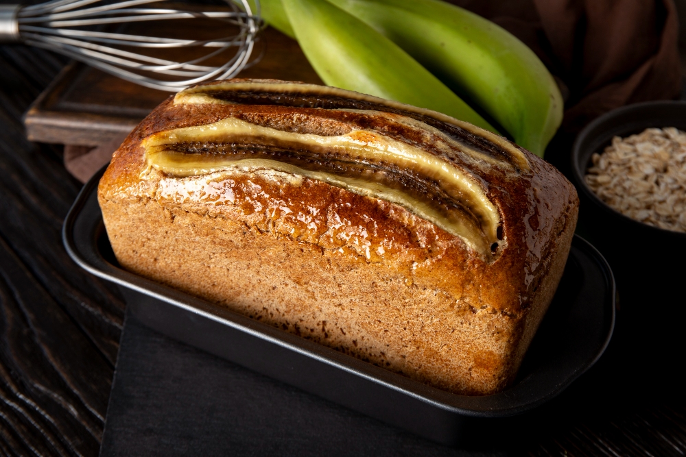 Freshly baked banana bread in a loaf pan with a golden crust, topped with sliced bananas, set on a dark wooden surface with green bananas and oats in the background.