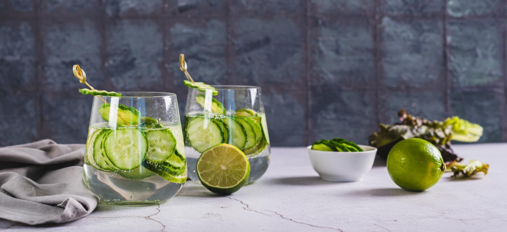 Two glasses of Hugo Spritz garnished with cucumber slices, served with fresh limes on a light gray table, perfect for summer refreshment.