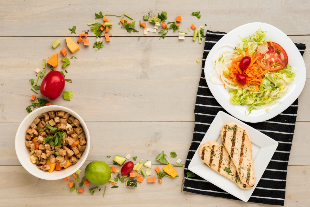 A healthy lunch setup featuring grilled chicken breast, fresh vegetable salad, and a bowl of tofu stir-fry on a wooden table with vibrant ingredients.