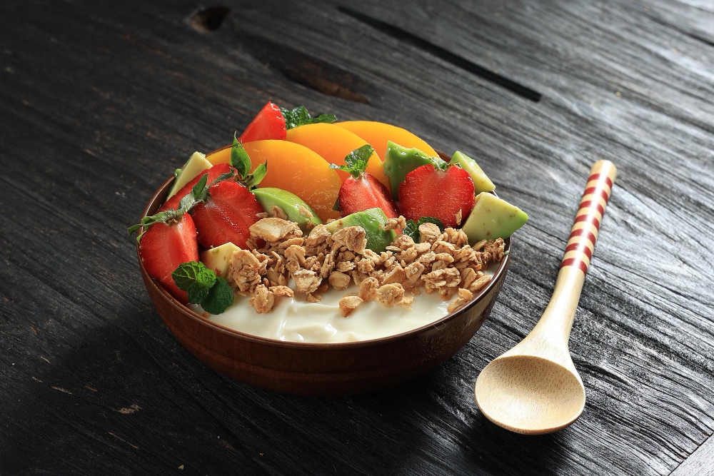 Healthy yogurt bowl with fresh fruits like strawberries, peaches, and avocado, topped with granola and mint leaves.