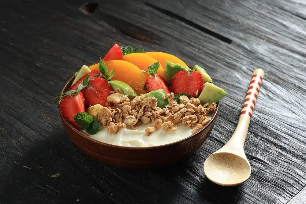 Healthy yogurt bowl with fresh fruits like strawberries, peaches, and avocado, topped with granola and mint leaves.