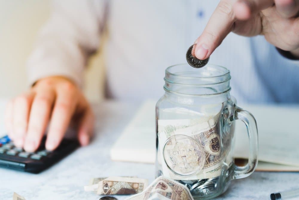 A person saving money in a glass jar while using a calculator, symbolizing budgeting and financial planning.