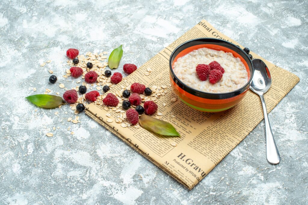 A bowl of oatmeal topped with fresh raspberries and blueberries, with scattered oats and a spoon on a rustic newspaper background.