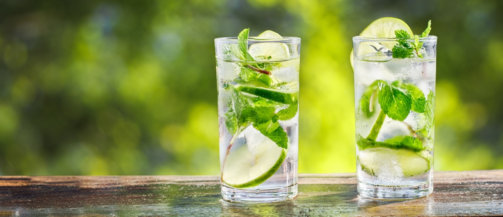 Two glasses of Hugo Spritz cocktail garnished with fresh mint and lime slices, placed on a wooden table with a blurred green background.