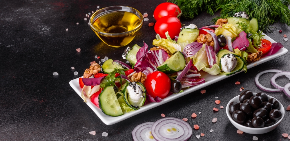 Low-carb salad with fresh vegetables, cucumbers, cherry tomatoes, black olives, walnuts, and cream cheese on a white rectangular plate