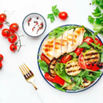 A healthy gluten-free meal on a white table featuring grilled chicken, fresh cherry tomatoes, mixed greens, grilled vegetables, and parsley.