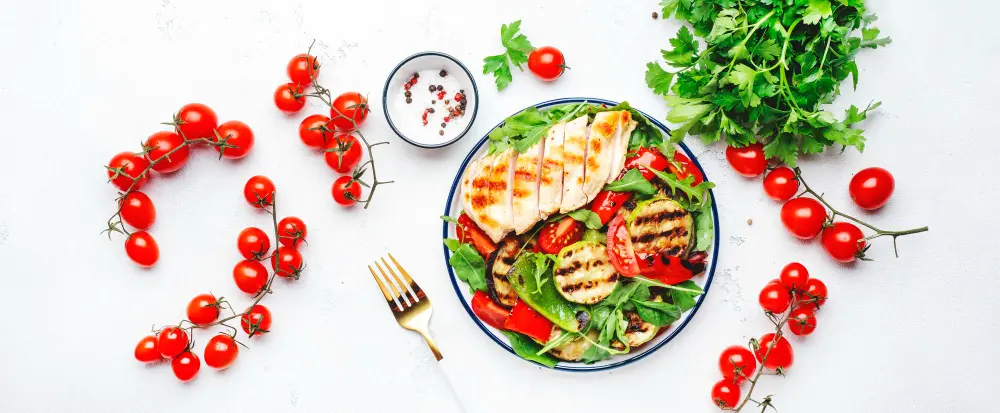 A healthy gluten-free meal on a white table featuring grilled chicken, fresh cherry tomatoes, mixed greens, grilled vegetables, and parsley.