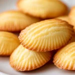 A close-up of golden, buttery creamy madeleine cookies arranged on a white plate, showcasing their delicate shell-like shape and soft texture. Perfect for breakfast